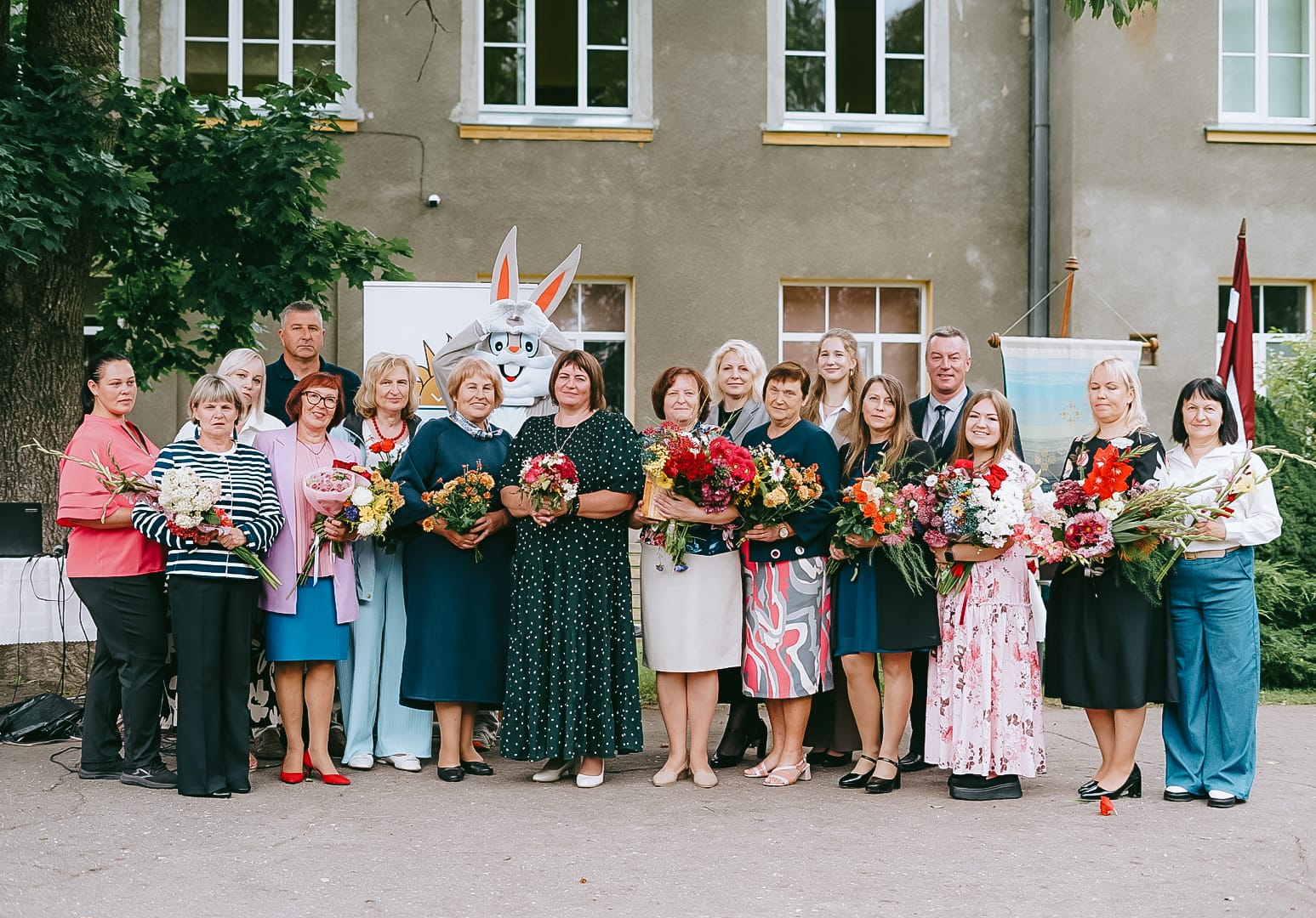 Vecsaules pamatskolas pedagogi un skolas darbinieki ar ziedu pušķiem Zinību dienas pasākumā pie skolas ēkas. Grupas fotoattēlā redzami skolas darbinieki, bet fonā – skolas ēka, Latvijas karogs un svinību noformējums.